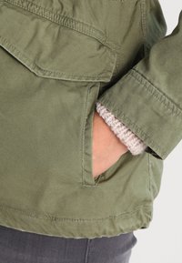 Grüne Canvas-Jacke mit einer aufgesetzten Tasche und einem strukturierten grauen Strickbündchen. Zeigt eine lockere Passform und sichtbare Nähte.