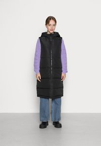 Gilet rembourré noir avec capuche, doté d'une fermeture éclair à l'avant, associé à un pull en tricot torsadé violet clair et à un jean large bleu.