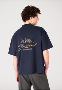 Camiseta de algodón azul marino de mangas cortas, con un diseño de escritura dorada y rama de olivo en la parte posterior, con el texto "Prohibido 10119 BERLÍN."