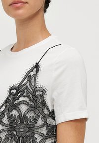 Balta trumpa rankovė t-shirt marškiniai su išsamiais juodais nėrinių akcentais priekyje, su suverstomis rankovėmis ir apvaliu iškirpimu.