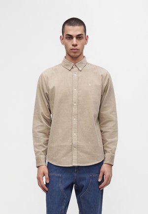 Jeune homme aux cheveux courts portant une chemise en velours côtelé beige boutonnée et un jean bleu, debout devant un fond clair uni.