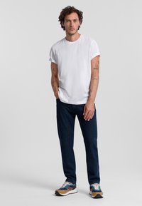 Witte katoenen T-shirt met opgerolde mouwen, donkerblauwe jeans en multifunctionele sneakers in meerdere kleuren. De persoon staat met een neutrale uitstraling tegen een effen achtergrond.