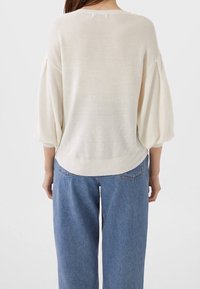 Suéter de punto ribeteado color crema con mangas anchas tipo globo, hombros caídos y un dobladillo que se curva ligeramente en la parte trasera. Combinado con jeans azules.