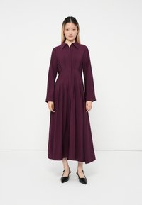 Frau, die vor einem weißen Hintergrund steht, trägt ein langärmliges, tiefpurpurfarbenes Knöchellanges Kleid und schwarze spitz zulaufende Schuhe.