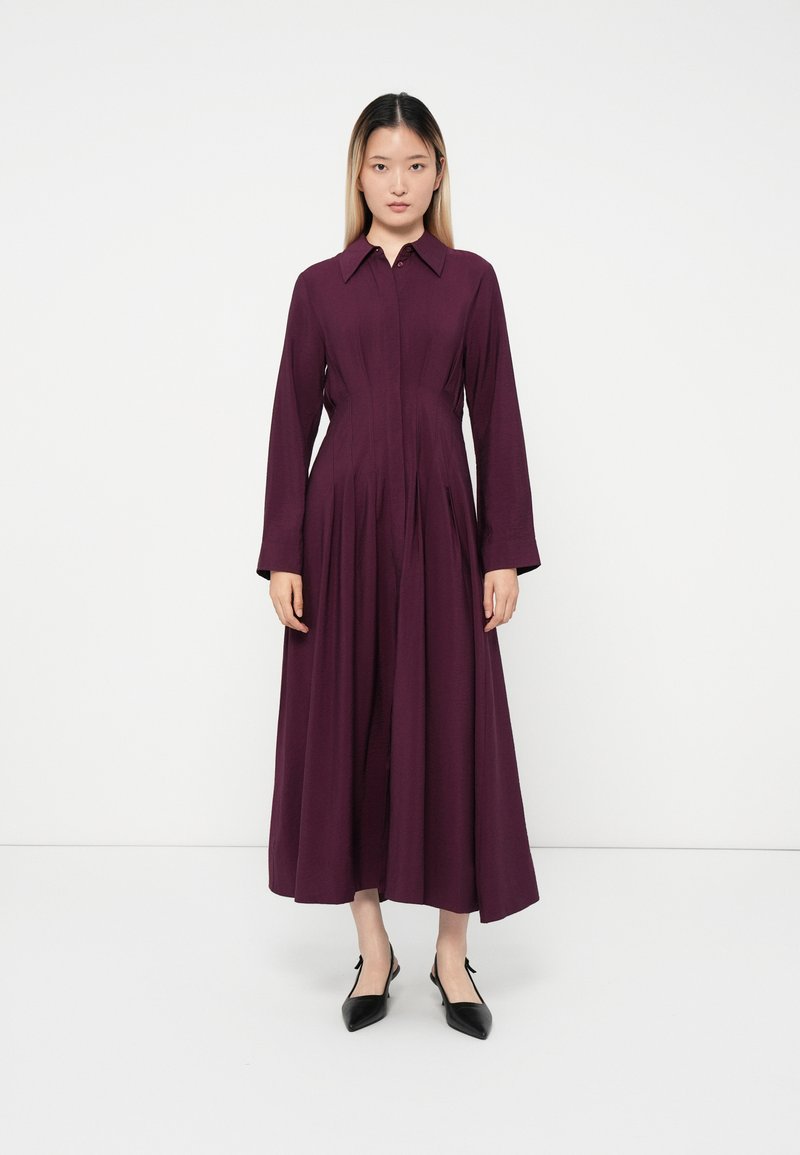 Frau, die vor einem weißen Hintergrund steht, trägt ein langärmliges, tiefpurpurfarbenes Knöchellanges Kleid und schwarze spitz zulaufende Schuhe.