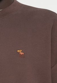 Καφέ πλεκτό πουλόβερ με στρογγυλή λαιμόκοψη και μικρό κεντημένο σχέδιο από άλκη σε καφέ και κίτρινο στο στήθος.