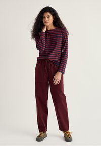 Csíkos, hosszú ujjú felső sötétkék és piros színekben, lazán szabott bordó nadrággal párosítva. A lábbeli olíva zöld színű, narancssárga fűzőkkel.