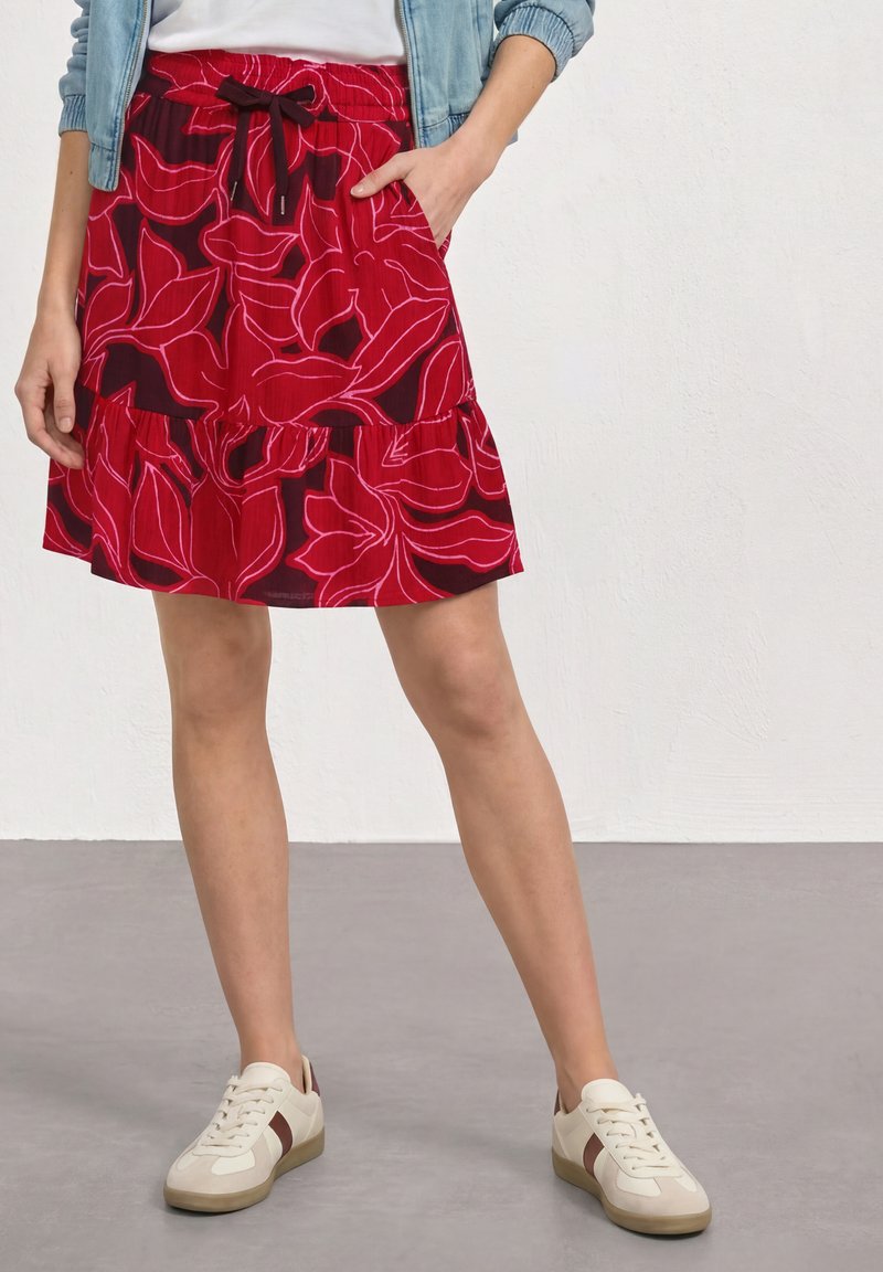 Femme portant une jupe rouge à fleurs avec une taille à cordon, des baskets blanches et une veste en jean claire, debout avec une main dans la poche.