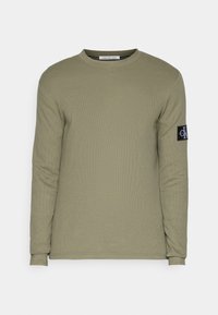 T-shirt à manches longues vert olive en tissu texturé. Présente un col rond et un patch noir avec un logo sur la manche gauche.