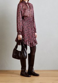 Donna in un vestito a maniche lunghe con fantasia floreale, che tiene una borsa a tracolla di pelle nera, indossando collant neri e stivali alti fino al ginocchio in pelle nera.
