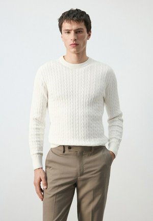 Joven de pie con un suéter blanco de punto grueso y pantalones beige, con una mano en el bolsillo, frente a un fondo liso.