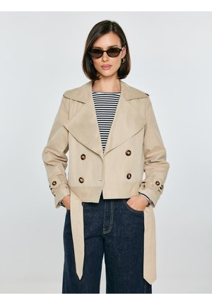 Moteris su akiniais nuo saulės, dėvinti smėlio spalvos dvieiliškį trumpą tranšėjos paltą, dryžuotą marškinius ir tamsius platėjancius džinsus, rankos kišenėse.