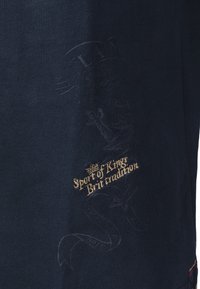 Ciemnogranatowa tkanina z delikatnym emblematem i złotym haftowanym napisem "Sport Królów Brytyjska tradycja" blisko środka.