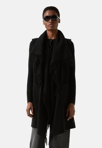 Sort oversized sjal med frynsete kanter, lagdelt over en sort top. Har lommer, båret med mørke jeans og runde solbriller.