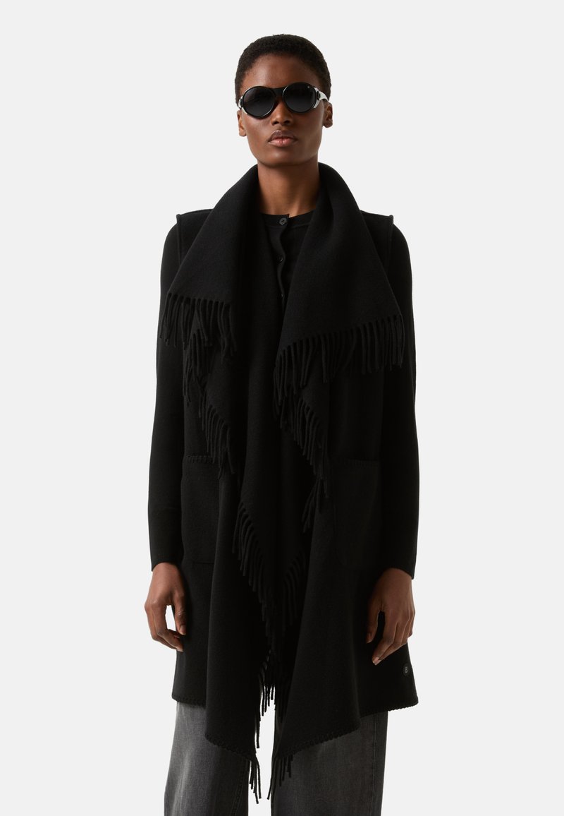 Sort oversized sjal med frynsete kanter, lagdelt over en sort top. Har lommer, båret med mørke jeans og runde solbriller.