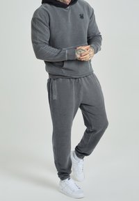 Grijze fleece hoodie met een zwarte capuchon, bijpassende grijze fleece joggers en witte sneakers. Heeft een logo op de borst en zijzakken.
