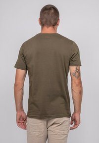 T-shirt kaki vert à manches courtes avec un col rond et une texture lisse, vu de dos. Présente un tatouage sur le bras gauche.
