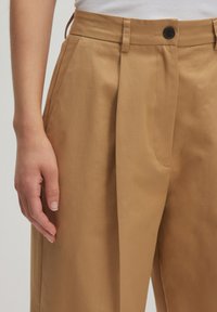 Tan geplooide broek gemaakt van een gladde stof, met een knoopsluiting, lussen voor een riem en zijzakken. Close-up van de tailleband en hand.