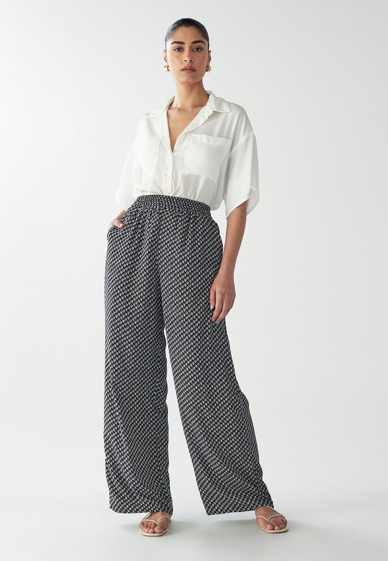Camicetta bianca a maniche corte con taschino; pantaloni larghi in bianco e nero con motivo e cintura elastica; modello che indossa sandali bassi.