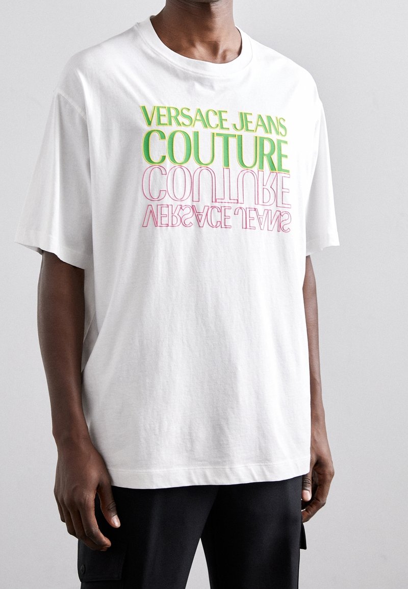 Bílý bavlněný tričko s multicolor textem "VERSACE JEANS COUTURE" v zelené a růžové barvě, volný střih s krátkým rukávem.