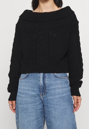 Sort, strikket crop-sweater med bred, ribbet off-shoulder krave og kabeldetaljer foran. Sættes sammen med high-waisted lyseblå jeans.