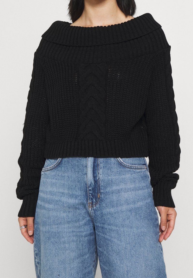 Black, knitted crop sweater with wide, ribbed off-shoulder collar and cable detailing on the front. Paired with high-waisted light blue jeans.