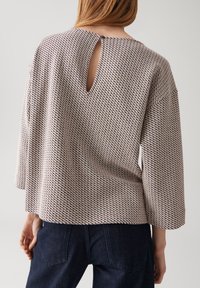 Femme portant un top beige ample à motifs avec une ouverture en forme de clé au dos et un bouton, associé à un jean bleu foncé, de dos.