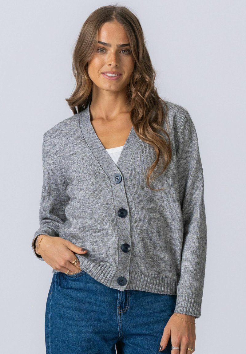 Femme portant un cardigan gris boutonné par-dessus un haut blanc et un jean bleu, souriante avec des cheveux bruns ondulés sur un fond uni.