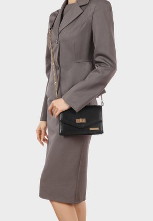 Donna che indossa un completo grigio, tiene una borsa nera in pelle con tracolla a catena dorata e chiusura in metallo, in piedi davanti a uno sfondo semplice.