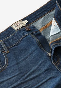 Blauwe denim jeans met een bronzen knoop en rits, met oranje stiksel en een label. De stof heeft een soepele textuur en een klassieke rechte snit.