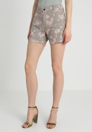 Kvinde iført beige blomstrede shorts med knap og lynlås, kombineret med metallisk grønne højhælede sandaler med ankelrem.