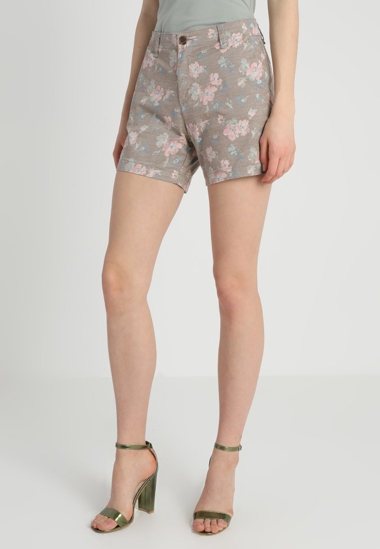 Femme portant un short beige à imprimé floral avec bouton et fermeture éclair, assorti à des sandales à talons hauts vert métallisé avec bride à la cheville.