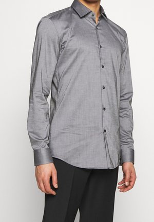 Homme portant une chemise grise à manches longues avec des boutons noirs, associée à un pantalon noir, debout devant un fond uni.