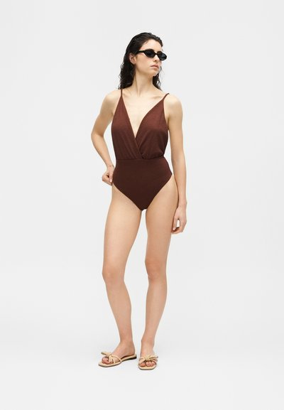 Femme portant un maillot de bain une pièce marron foncé texturé, des lunettes de soleil noires et des sandales beige à nœud, debout avec une main sur la hanche, sur fond blanc.