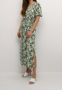 Floral Maxi-Kleid in Grün, Weiß und Blau mit V-Ausschnitt und kurzen Ärmeln. Verfügt über seitliche Schlitze und wird mit weißen High Heels kombiniert.