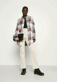 Camisa xadrez em cinza e castanho, blusa preta por baixo, calças brancas de cintura alta, botas pretas, segurando uma bolsa preta texturizada.