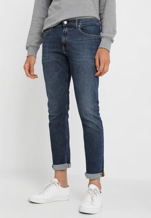 Person iført mørkeblå, opsmøgede jeans, hvide sneakers og en grå langærmet skjorte, stående mod en ensfarvet baggrund.