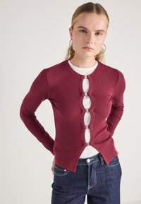 Cardigan ribbed burgundy, cu decolteu rotund, cu o serie de decupaje în formă de cheie și accente din metal argintiu pe partea frontală.