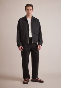 Black zip-up jacket with two front pockets, paired with black pants and brown sandals. The setting features a neutral background.