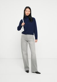 Donna che indossa un maglione a collo alto blu navy, jeans grigio chiaro, scarpe nere a punta, mentre tiene una piccola borsa bianca a tracolla, in piedi davanti a uno sfondo bianco.