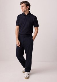 Temno modra polo majica s kratkimi rokavi in teksturiranim vzorcem, združena s temnimi hlačami in belimi supergami. Enostaven dizajn in krojen kroj.