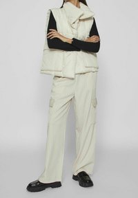 Vadderad, krämfärgad väst med vikkrage i kombination med krämrandiga vida byxor. Svart långärmad topp och svarta plattformsskor.