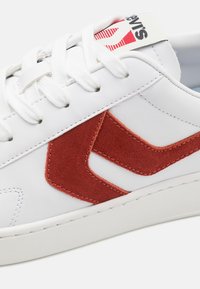 Białe sneakersy z gładkiej skórzanej powierzchni i czerwonymi zamszowymi akcentami. Posiadają płaskie białe sznurowadła i metkę z logo na języku.