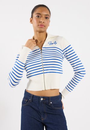 Cropped zip-up sweater in white with blue horizontal stripes and ribbed cuffs. Features "Smil" embroidered on the chest. Paired with dark jeans.
