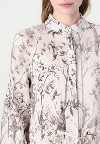 Bluza s cvetnim vzorcem iz svetlo kremnega materiala, ki ima podrobne botanične ilustracije v sivih odtenkih, z nabranim ovratnikom in pasom na vrvico.