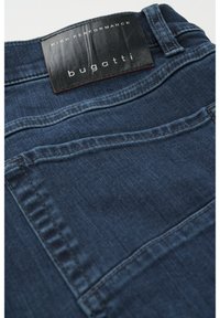 Mørkeblå denim jeans med en glat tekstur. Har et sort læder mærke med hvid tekst, placeret ved taljen.