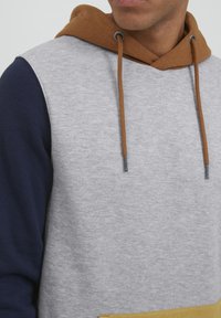 Grauer Sweatshirt mit navyblauen Ärmeln und einer braunen Kapuze. Verfügt über Kordeln und eine Fronttasche mit einem gelben Akzent. Weiche Textur.