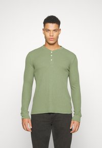 Zöld, hosszú ujjú Henley ing, bordázott anyagból készült, kerek nyakkivágással, elől négy gombbal és formára szabott ujjakkal.