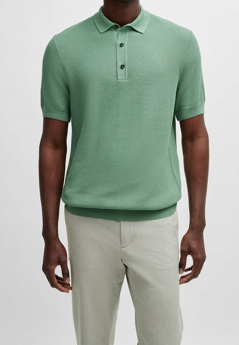 Homme portant un polo vert texturé à manches courtes et un pantalon beige clair, debout devant un fond blanc uni.