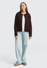 Brauner gerippter Cardigan mit rundem Ausschnitt und Knopfleiste, kombiniert mit einem weißen Oberteil und hellblauen weiten Jeans. Strukturiertes Material.